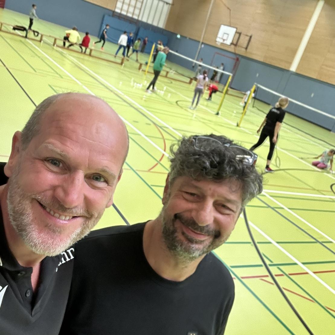 Zwei M&auml;nner l&auml;cheln in einer Sporthalle; im Hintergrund sind Kinder beim Badmintonspielen aktiv.