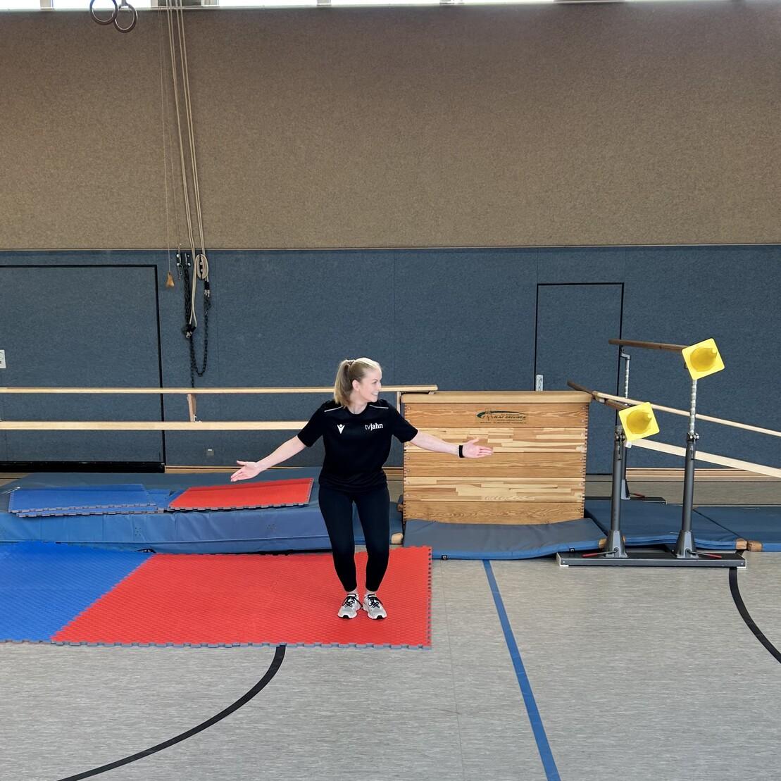 T&auml;nzerin in Schwarz auf einem Gymnastikboden, umgeben von Matten und einem Holzblock. Sie h&auml;lt die Arme ausgebreitet.