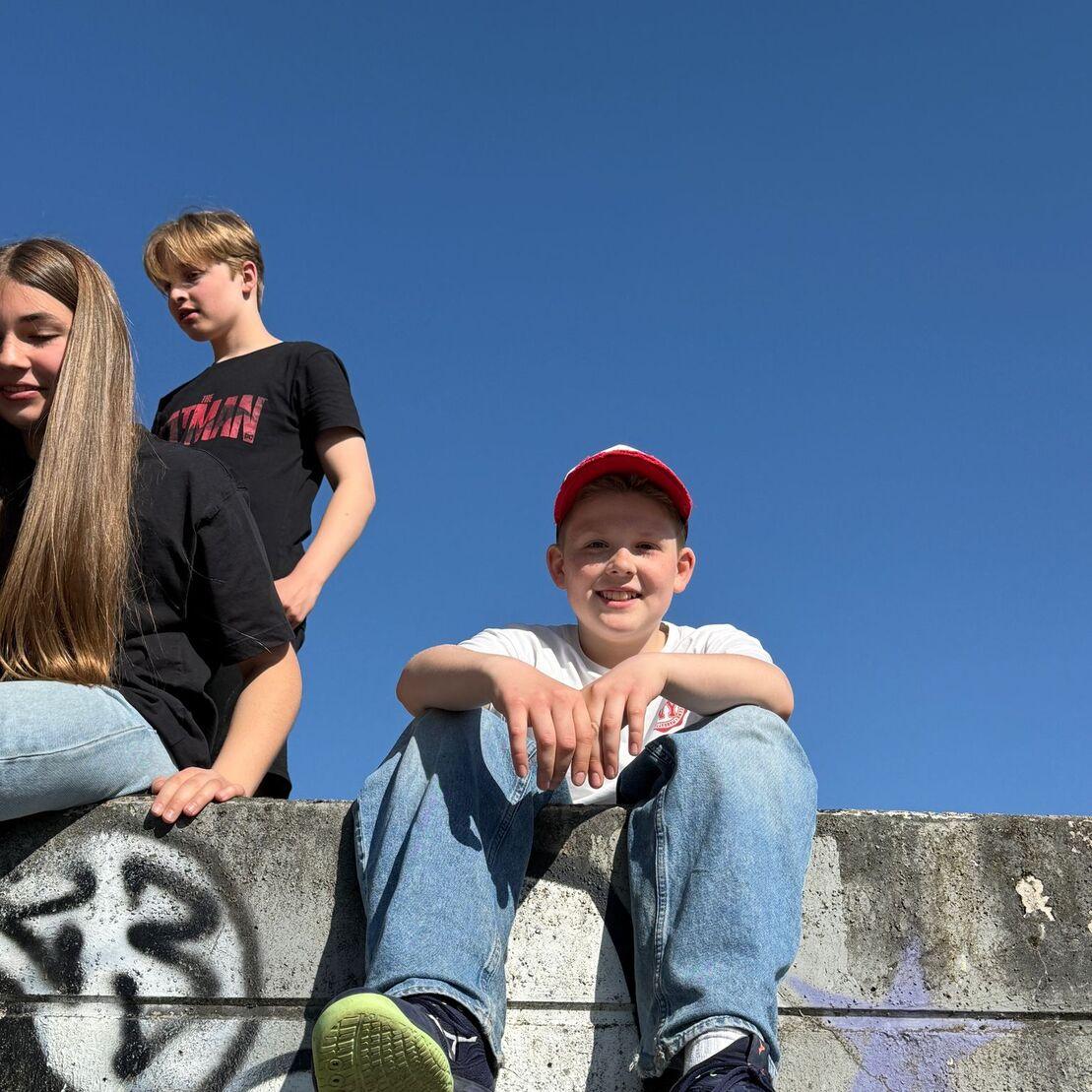 Drei Kinder sitzen auf einer Mauer unter blauem Himmel; ein Junge mit rotem Hut l&auml;chelt in die Kamera.