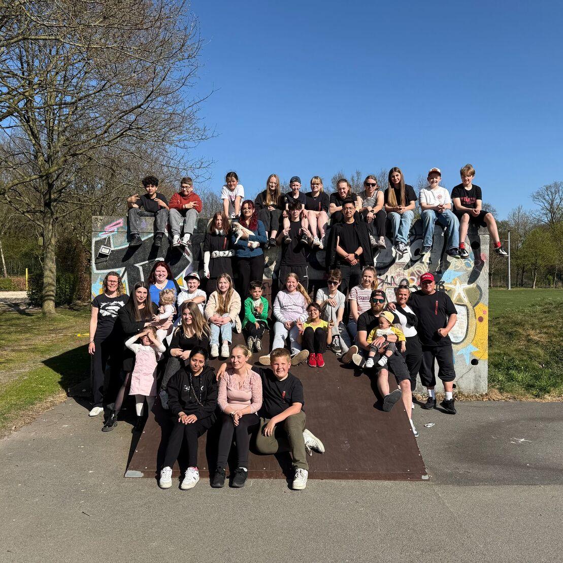 Gruppenfoto von etwa 30 Personen auf einer Skateboardrampe, umgeben von B&auml;umen und Wiese unter blauem Himmel.