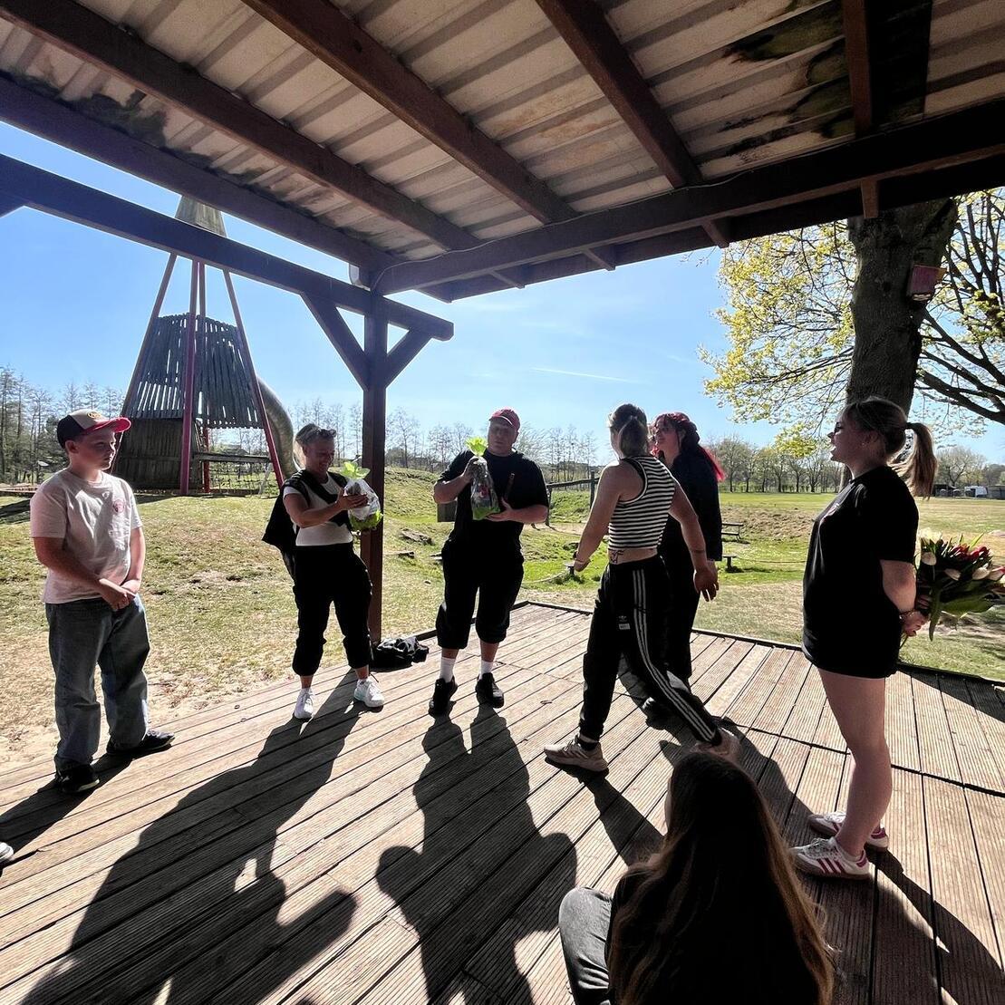Gruppe von Personen steht unter einem Holz&uuml;berstand und unterh&auml;lt sich bei sonnigem Wetter im Freien.