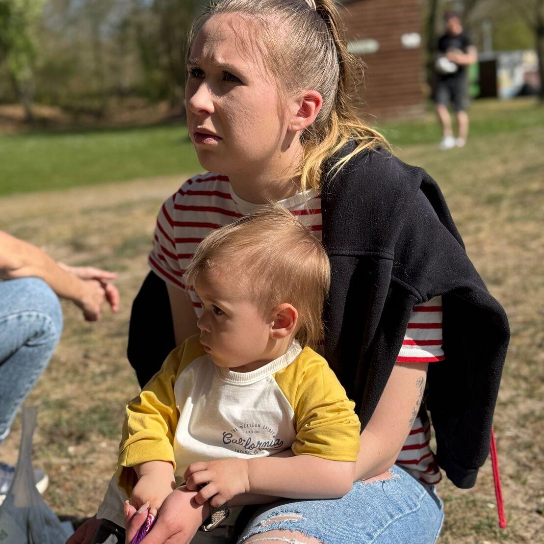 Junge Frau mit Zopf sitzt mit einem kleinen Kind auf dem Scho&szlig;. Beide schauen nachdenklich in die Ferne.