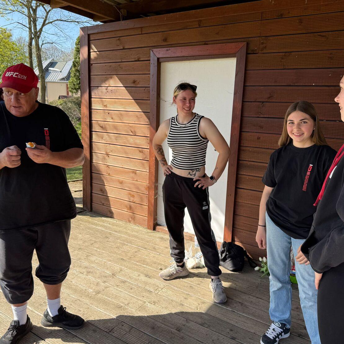 Vier Personen stehen in einer Holzlaube und unterhalten sich, w&auml;hrend ein Mann etwas in der Hand h&auml;lt.