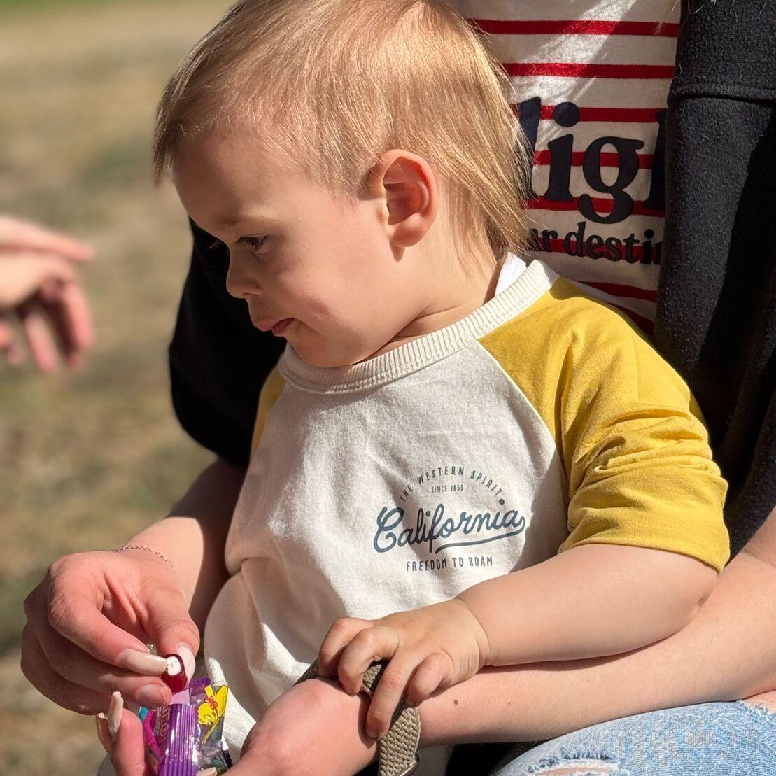 Kleines Kind mit blondem Haar sitzt auf dem Scho&szlig; einer Person und h&auml;lt eine T&uuml;te mit S&uuml;&szlig;igkeiten in der Hand.