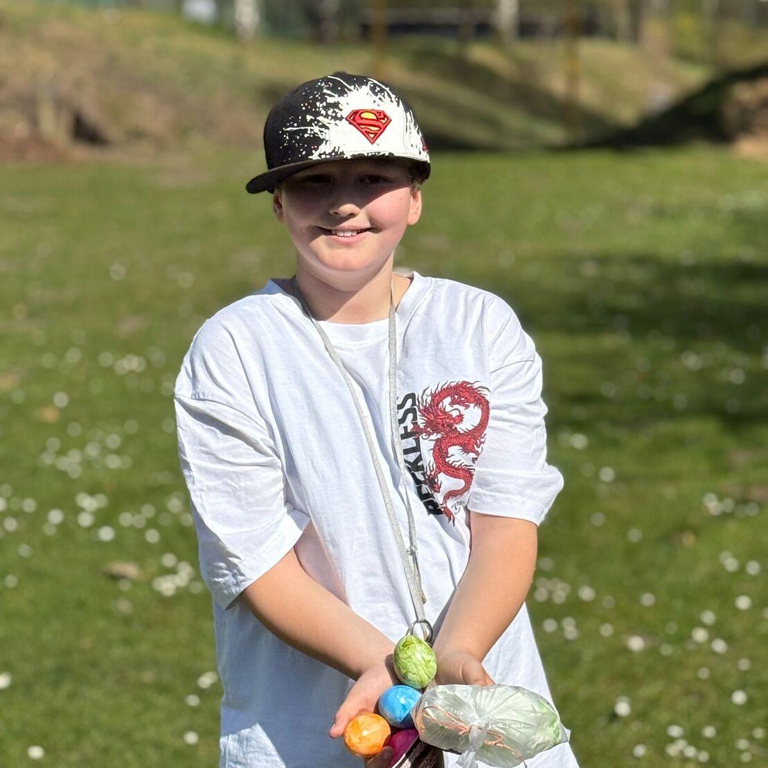 Ein Junge in einem wei&szlig;en T-Shirt und Cap h&auml;lt bunte Plastikb&auml;lle auf einem gr&uuml;nen Rasen mit B&auml;umen im Hintergrund.
