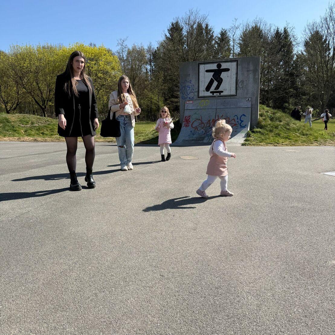 Eine kleine blonde Tochter l&auml;uft auf einem Gehweg, w&auml;hrend drei Frauen im Hintergrund stehen und zuschauen.
