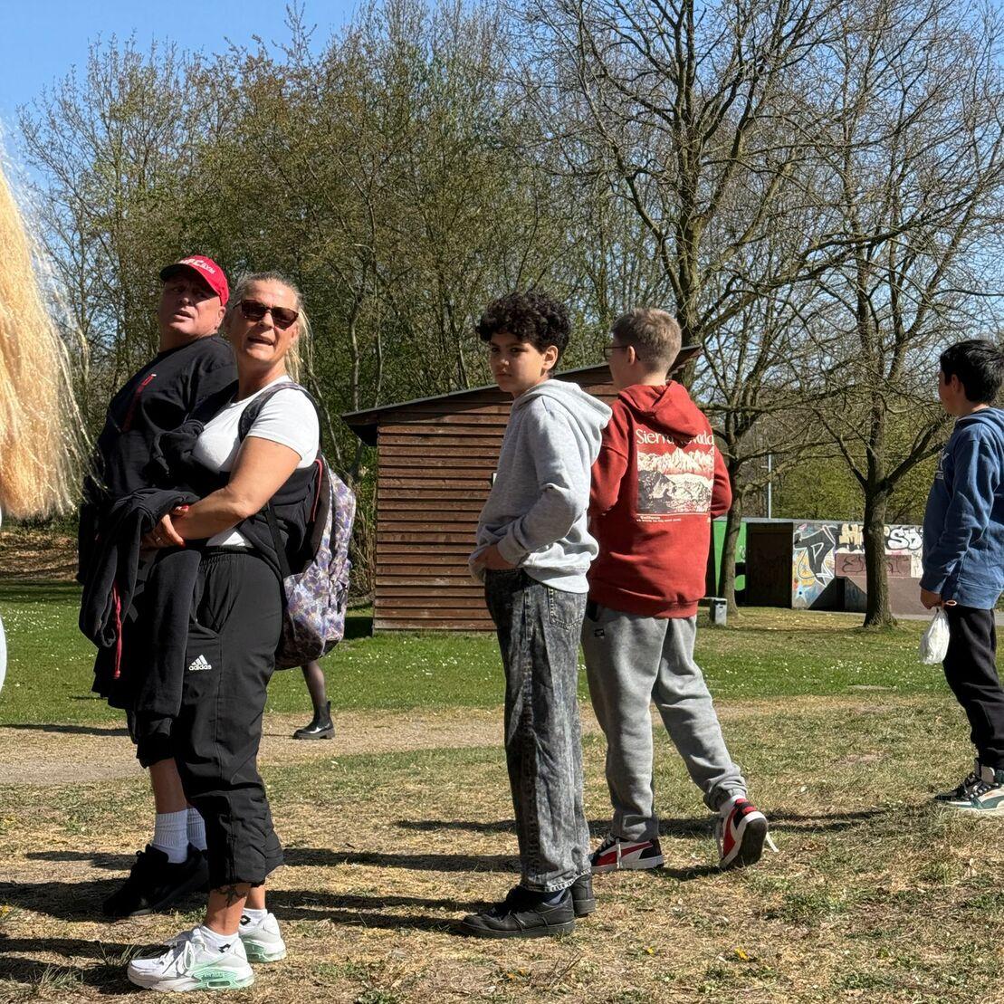 Gruppe von Kindern und Erwachsenen steht auf einer Wiese vor einem Holzschuppen, Sonnenlicht scheint durch die B&auml;ume.