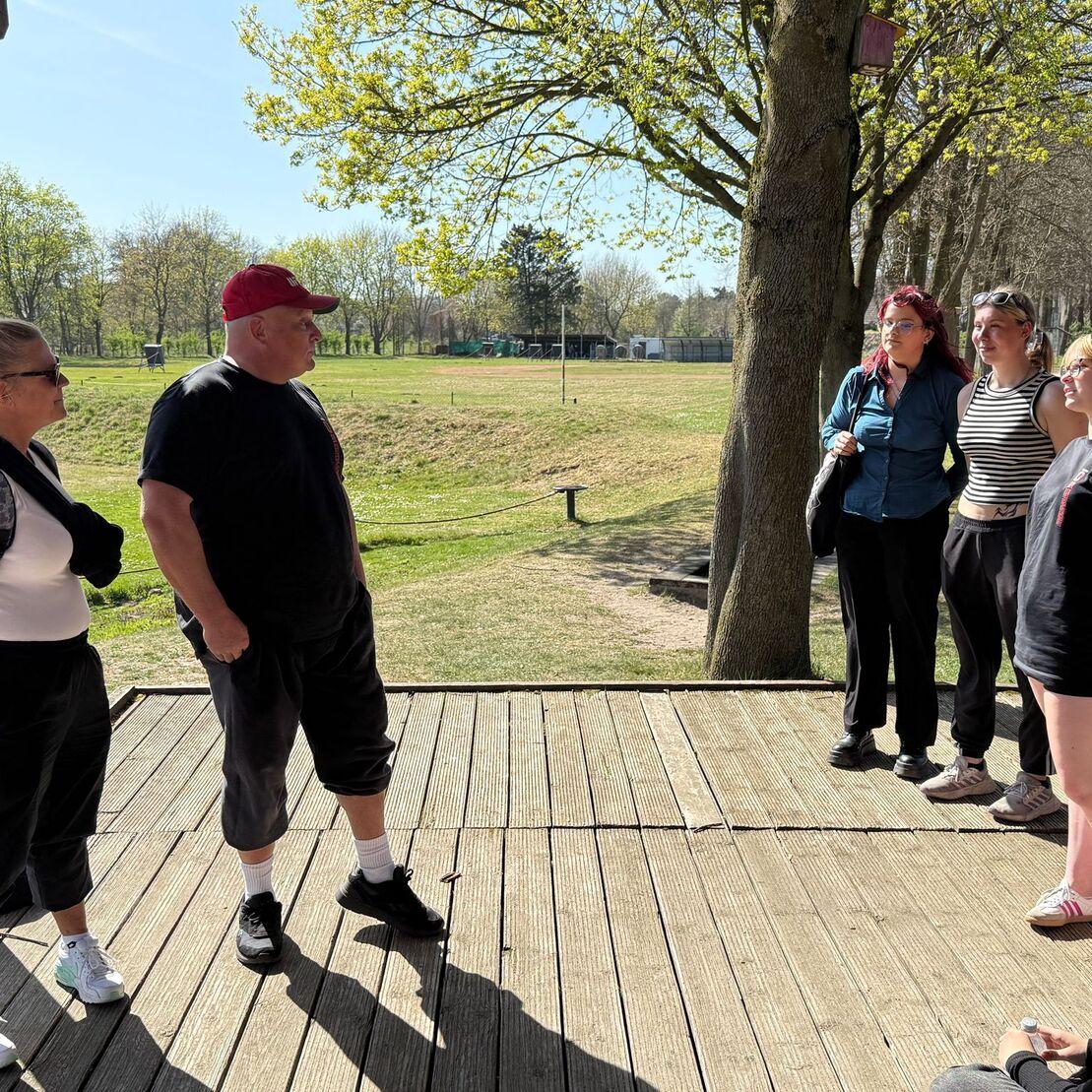 Sechs Personen stehen auf einer Holzterrasse unter einem Baum und sprechen miteinander, im Hintergrund eine gr&uuml;ne Wiese.
