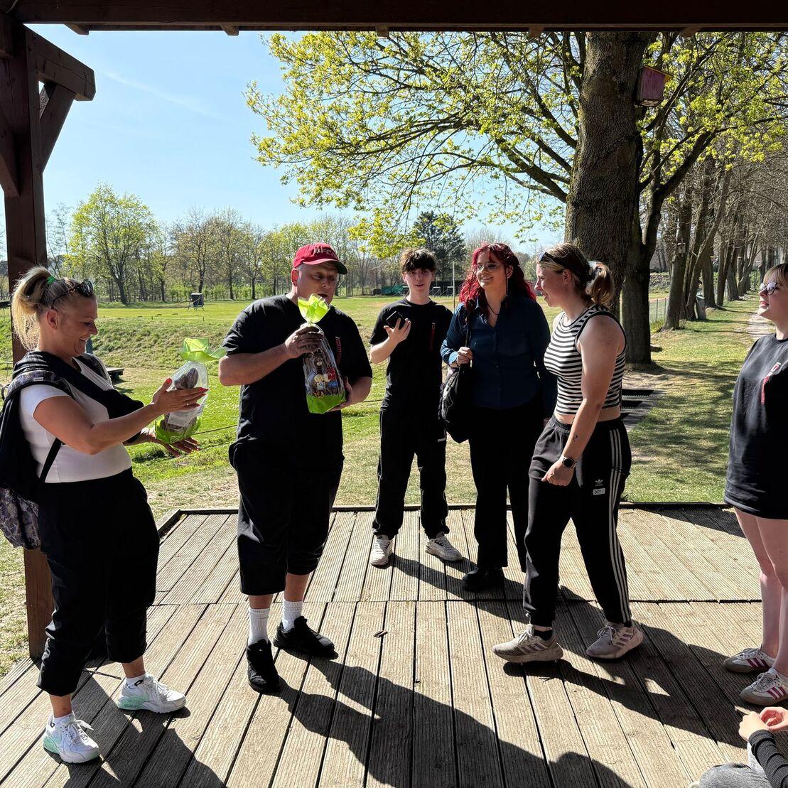 Gruppe von Personen auf einer Holzterrasse im Freien, einige interagieren und lachen, umgeben von B&auml;umen.