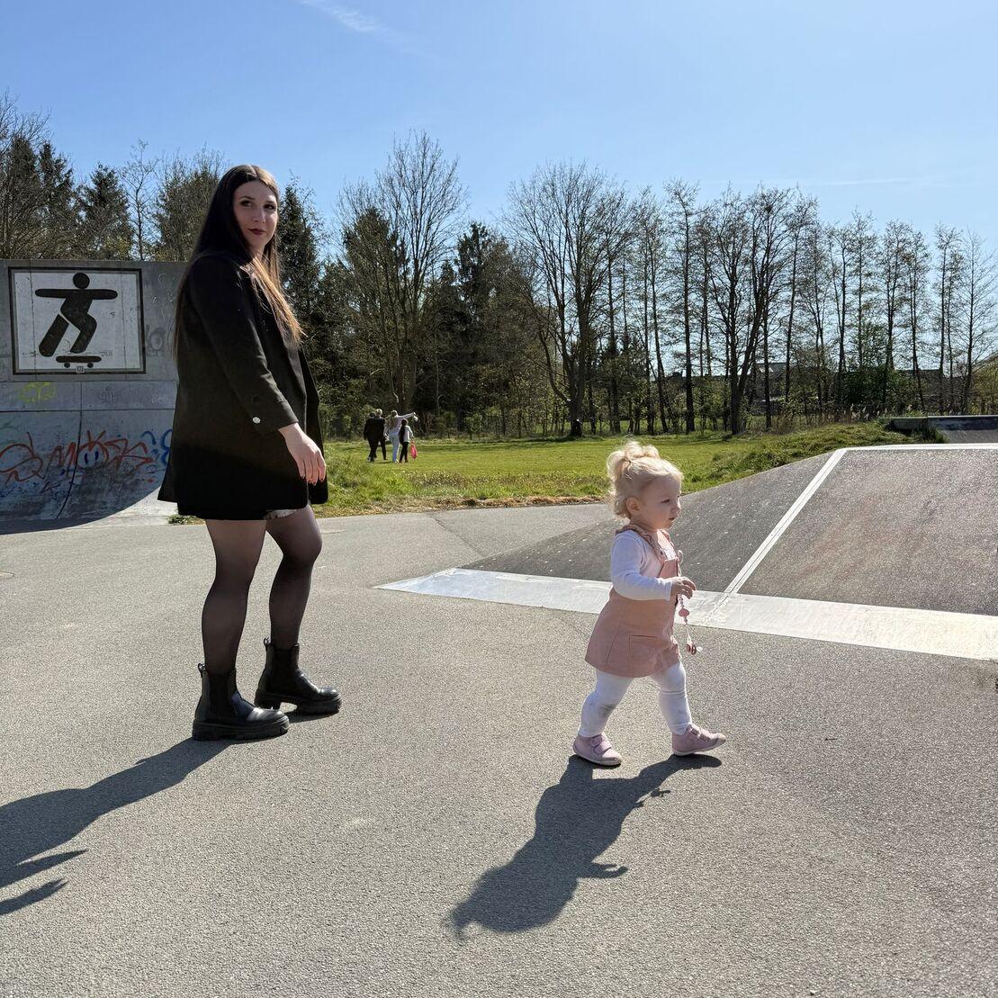 Frau mit langen Haaren steht neben einem kleinen M&auml;dchen auf einem Skateplatz an einem sonnigen Tag.