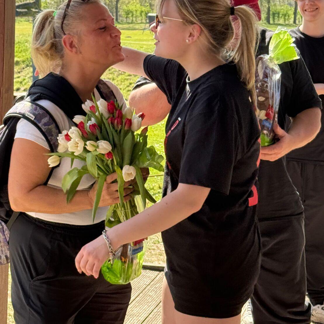 Ein M&auml;dchen &uuml;berreicht einer Frau mit Blumenstrau&szlig; in der Hand eine Umarmung im Freien. Im Hintergrund stehen weitere Personen.