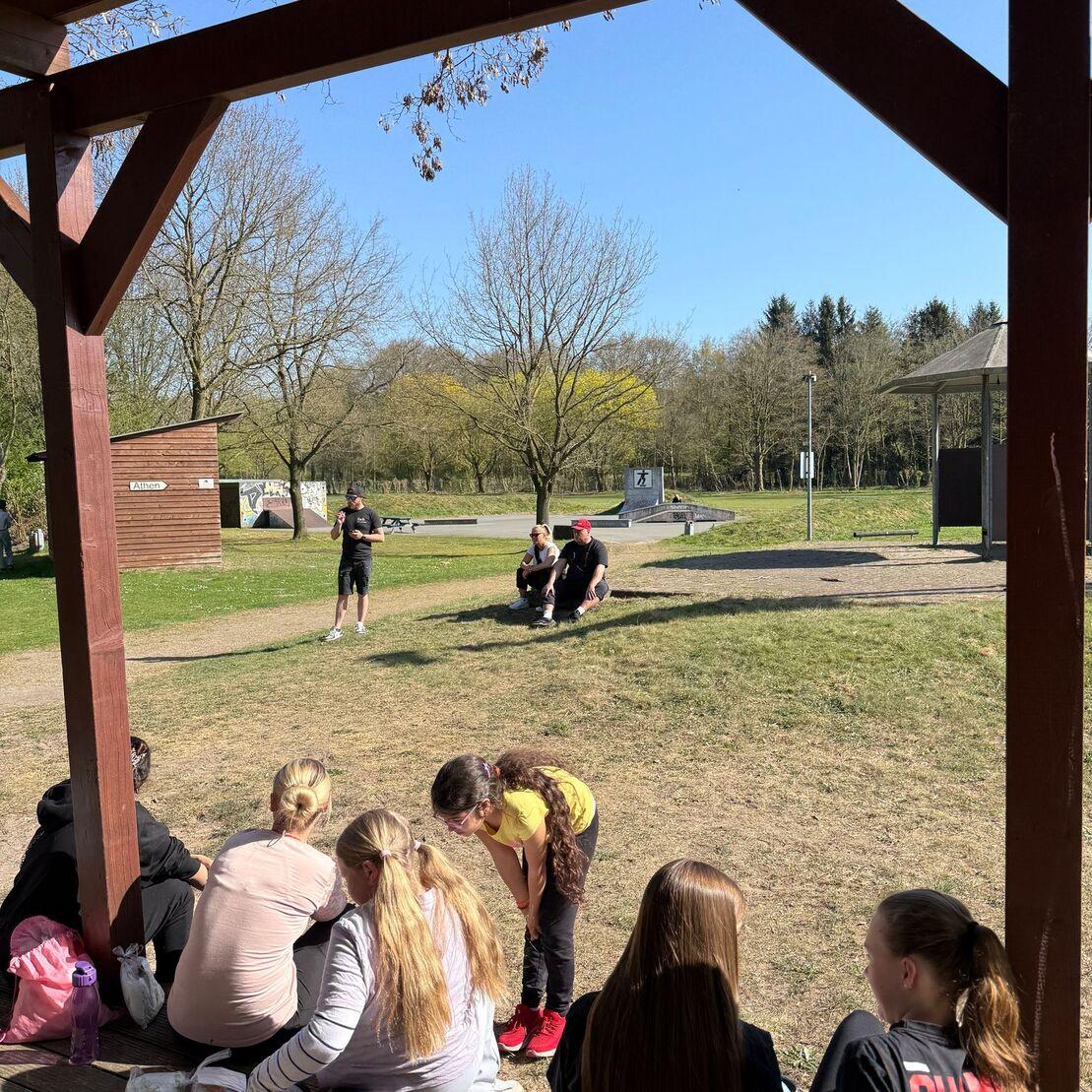 Gruppe von Kindern sitzt vor einem Pavillon, w&auml;hrend andere auf einer Wiese spielen und einen Aussichtspunkt beobachten.