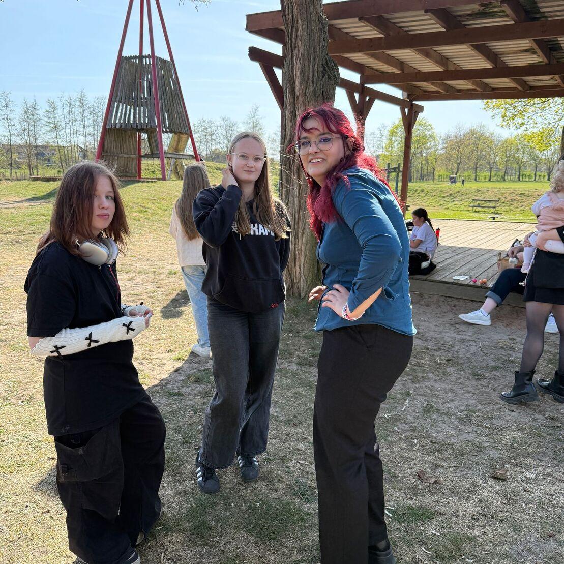 Drei junge Frauen stehen im Freien, vor einem Holzspielger&uuml;st, an einem sonnigen Tag.