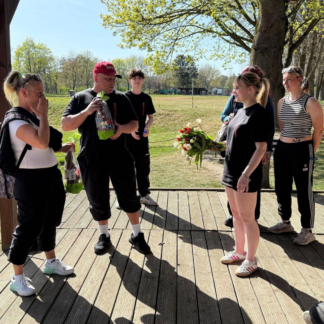 Gruppe junger Leute auf Holzplattform, einer h&auml;lt einen Blumenstrau&szlig;, sonniger Tag im Freien.