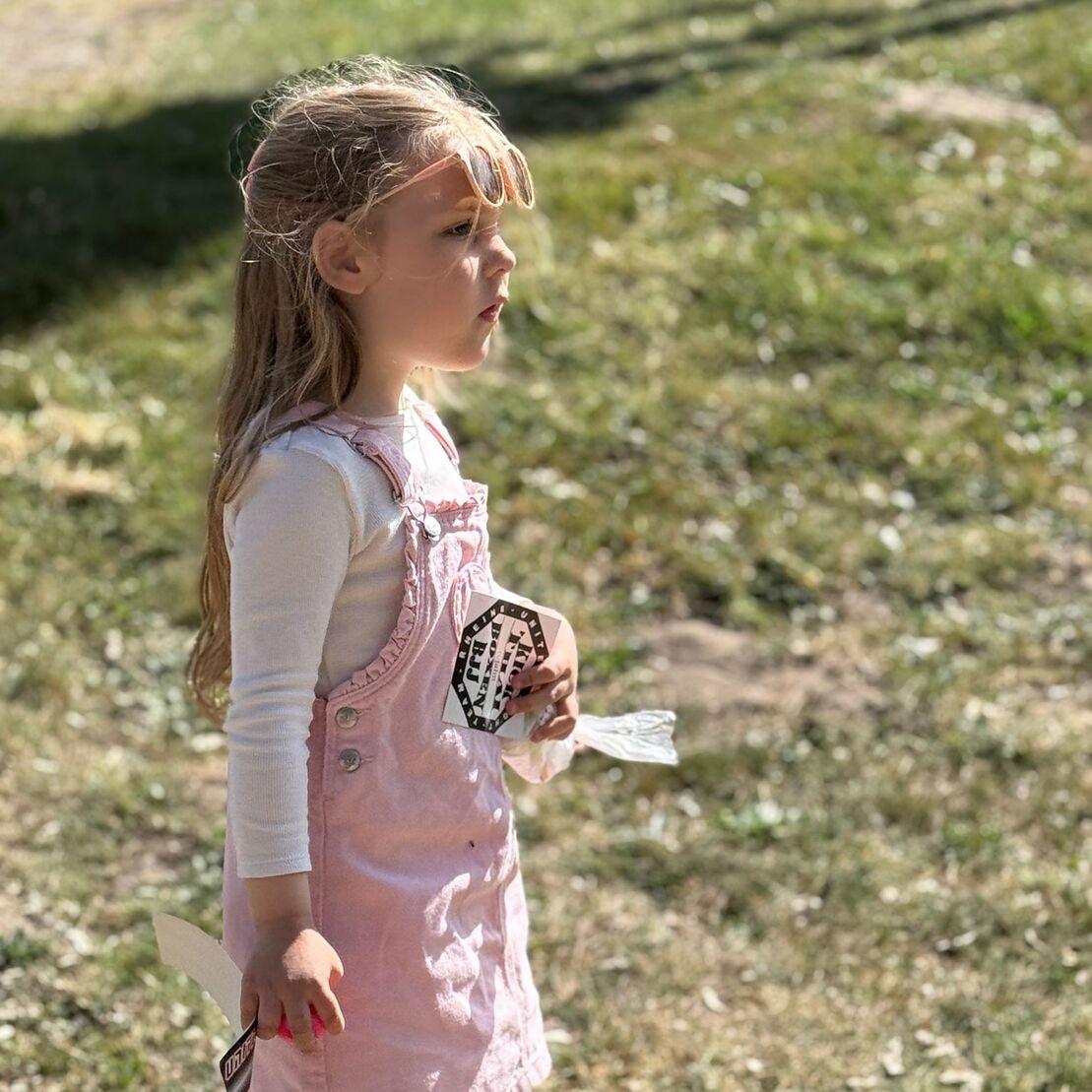M&auml;dchen mit langen Haaren in rosa Latzkleid geht auf einer gr&uuml;nen Wiese und h&auml;lt ein Papier in der Hand.