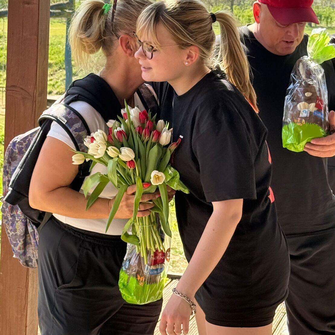 Zwei Frauen umarmen sich, eine h&auml;lt einen Strau&szlig; Tulpen, im Hintergrund ein Mann mit einer gr&uuml;nen T&uuml;te.