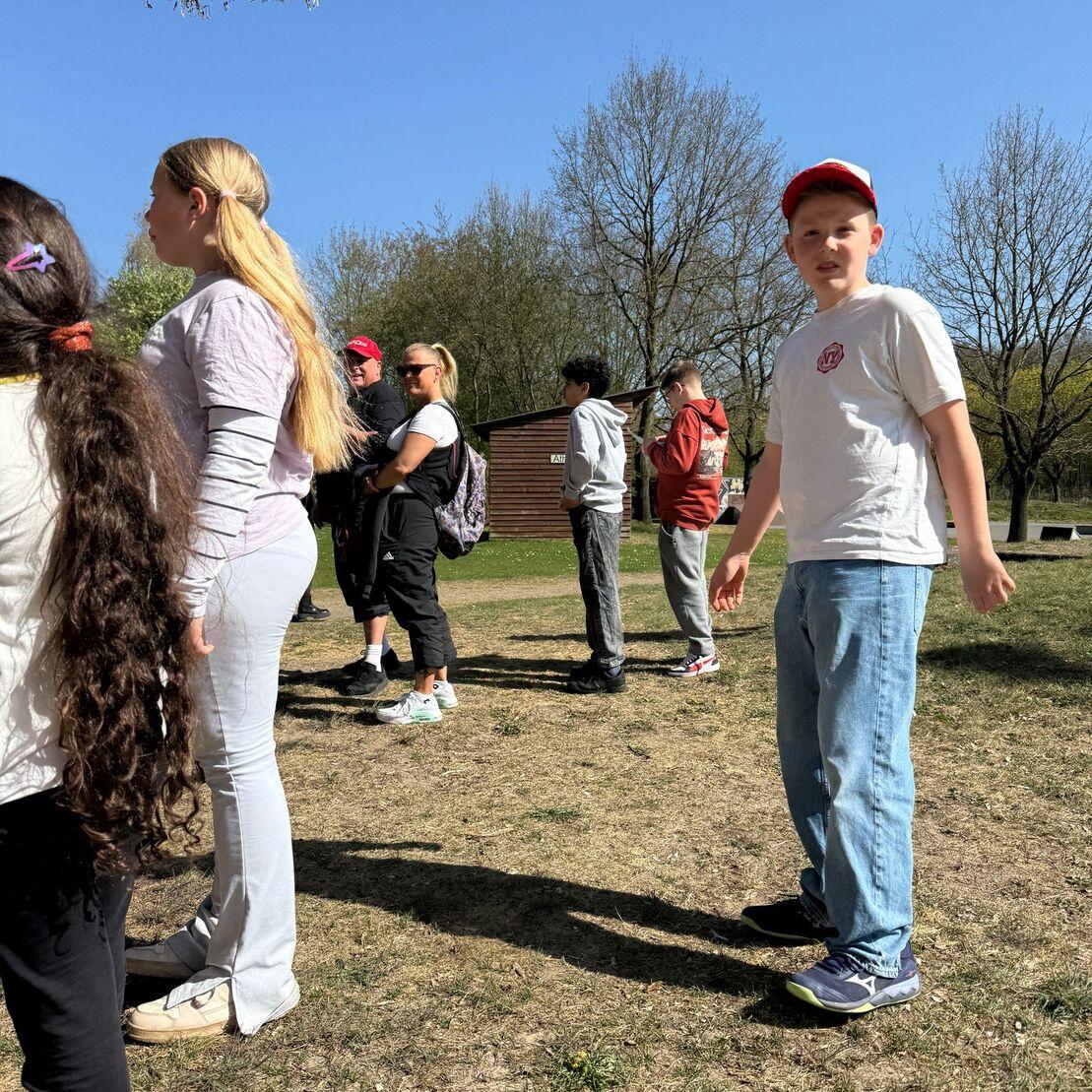 Gruppe von Kindern und Erwachsenen steht auf einer Wiese, umgeben von B&auml;umen und einem Holzgeb&auml;ude im Hintergrund.