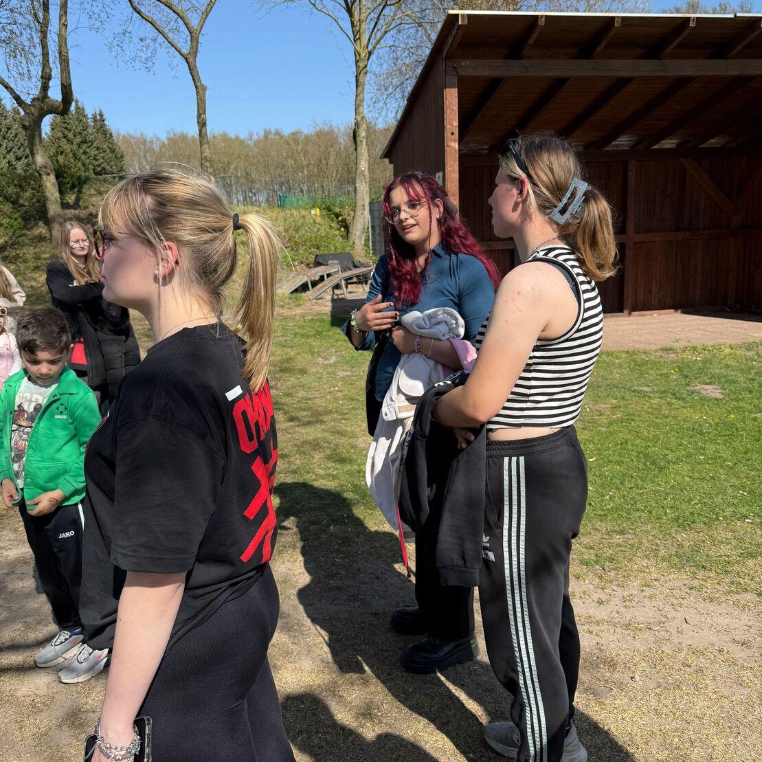 Gruppengespr&auml;ch im Freien: Jugendliche stehen auf einer Wiese, im Hintergrund eine Holzunterstand. Sonniger Tag.