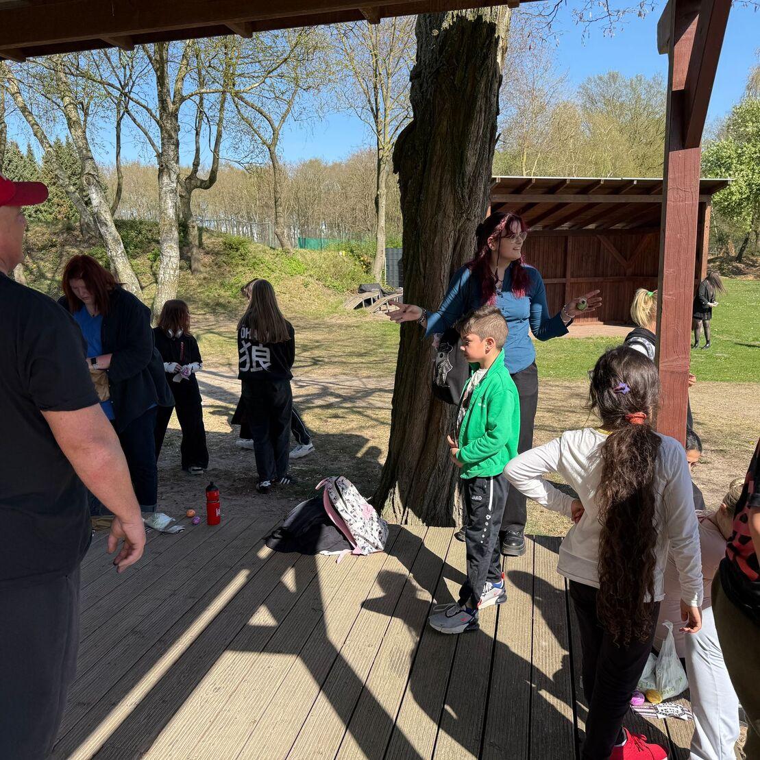 Gruppe von Kindern und Erwachsenen versammelt unter einem &uuml;berdachten Holzbereich in einer park&auml;hnlichen Umgebung.
