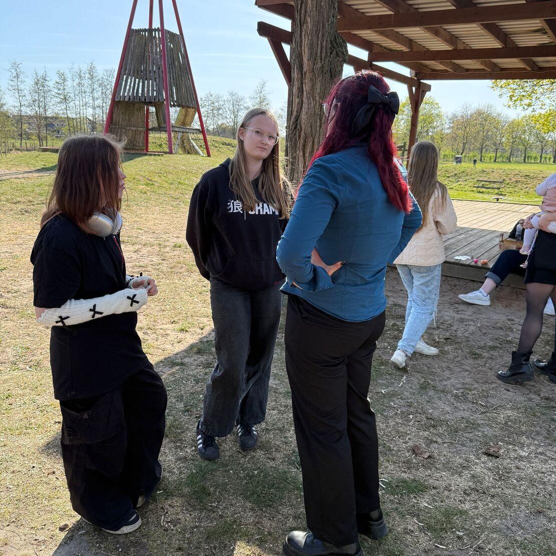 Drei Jugendliche unterhalten sich im Freien, im Hintergrund eine Spielstruktur aus Holz und B&auml;ume.