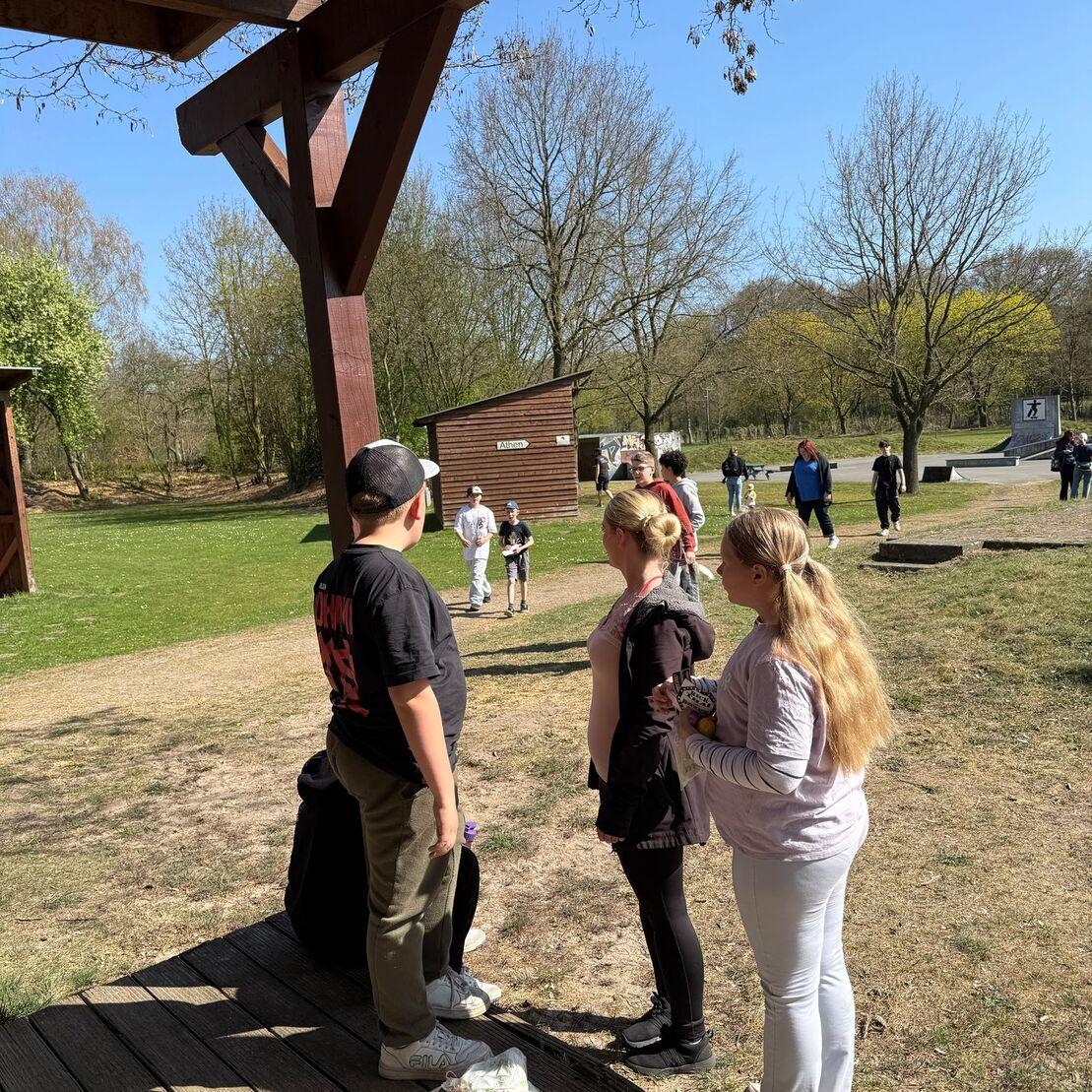 Drei Personen stehen auf einem Holzpodest, w&auml;hrend im Hintergrund eine Gruppe von Menschen im Park aktiv ist.
