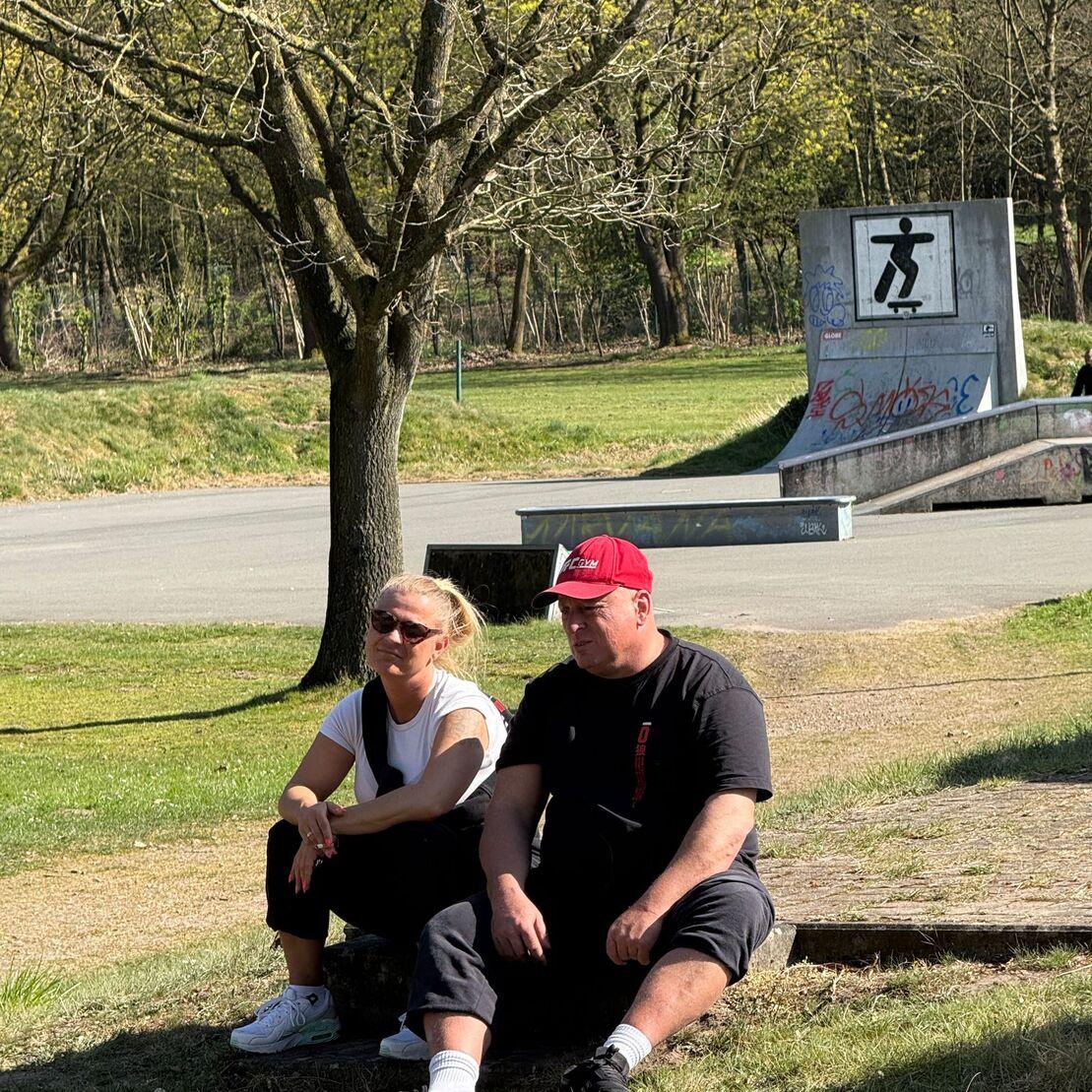 Zwei Personen sitzen auf dem Boden in einem Park, im Hintergrund eine Skateboardrampe und ein Wegweiser.