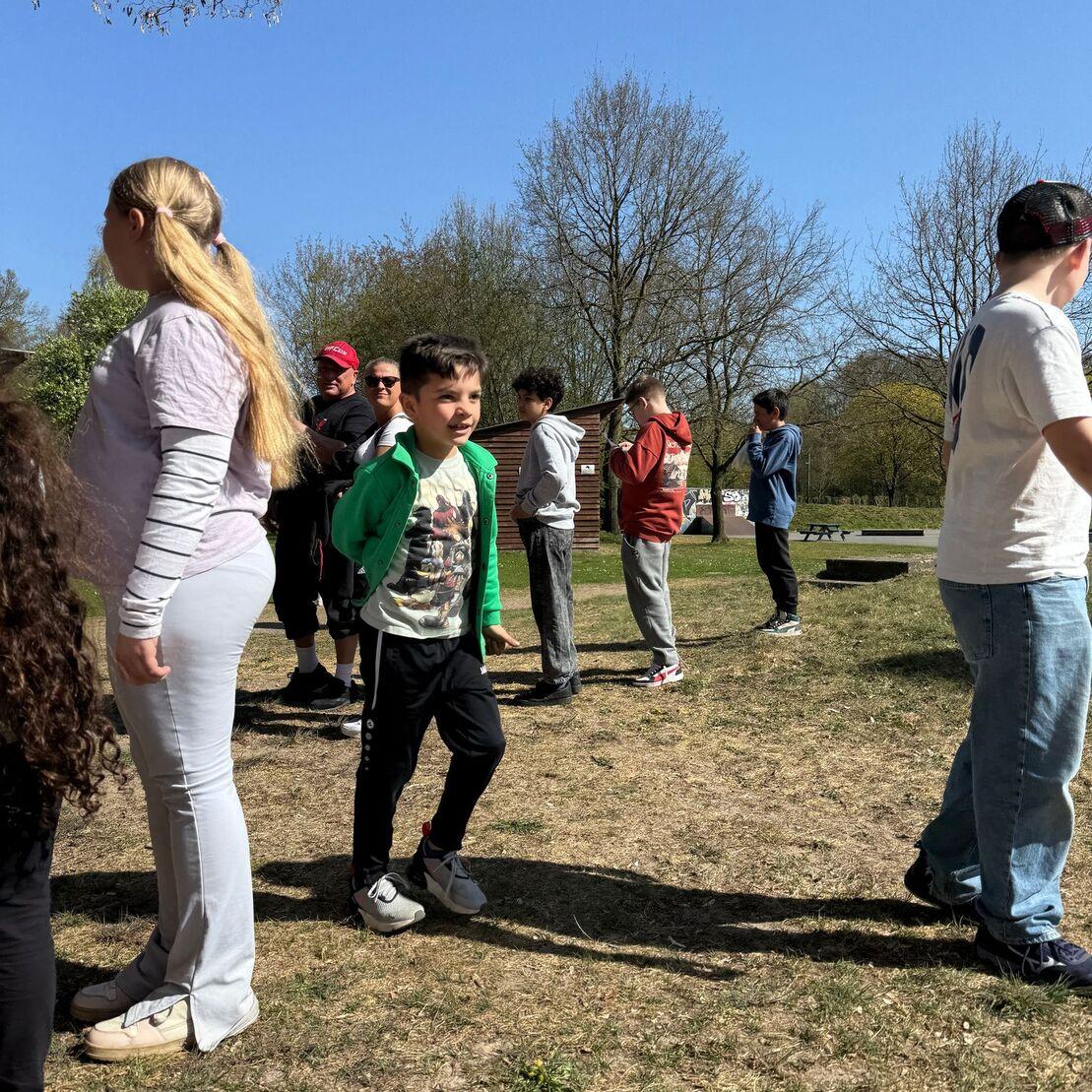 Gruppe von Kindern und Erwachsenen steht im Freien auf einer Wiese, umgeben von B&auml;umen und blauem Himmel.