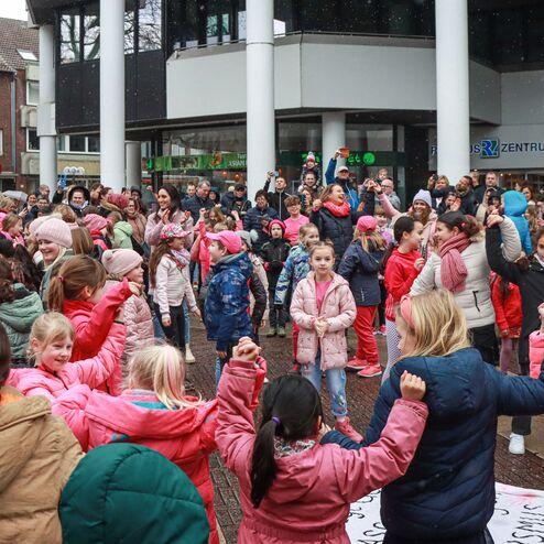 Gruppentanz von Kindern und Erwachsenen in bunten Winterjacken vor einem Geb&auml;ude, umgeben von Zuschauern.