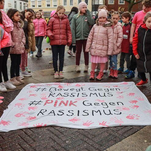 Gruppierung von Kindern in pinken Outfits um ein Banner mit der Aufschrift &bdquo;#Bewegt Gegen RASSISMUS&ldquo; auf dem Boden.