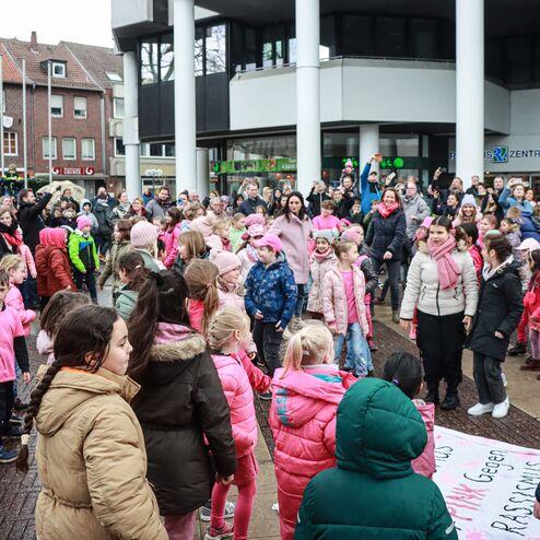 Gruppentanz von Kindern und Erwachsenen in pinken Outfits auf einer belebten Stra&szlig;e vor einem modernen Geb&auml;ude.