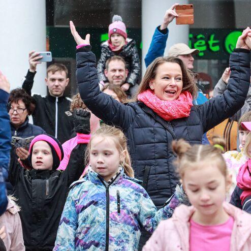 Gruppe von Menschen feiert drau&szlig;en, eine Frau in dunkler Jacke und rotem Schal hebt die Arme, umgeben von Kindern in bunten Jacken.