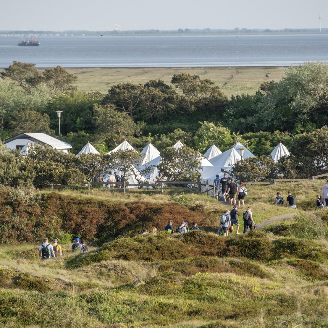 Blick auf eine K&uuml;stenlandschaft mit Gruppen von Menschen, Zeltanlagen und einem weiten Wasserhorizont im Hintergrund.