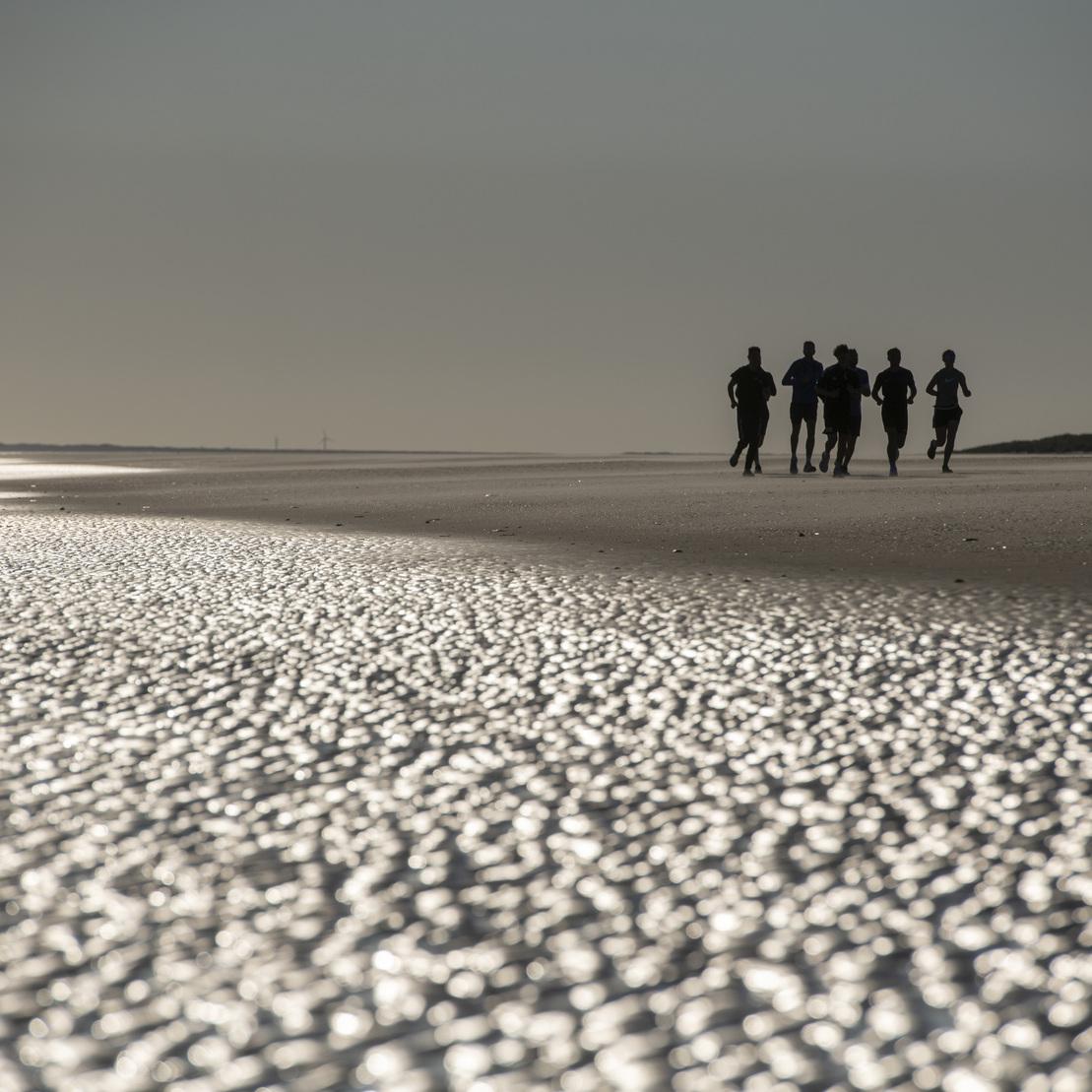 Gruppe von f&uuml;nf Personen geht am Strand &uuml;ber sandige Fl&auml;che, reflektierendes Licht im Hintergrund.