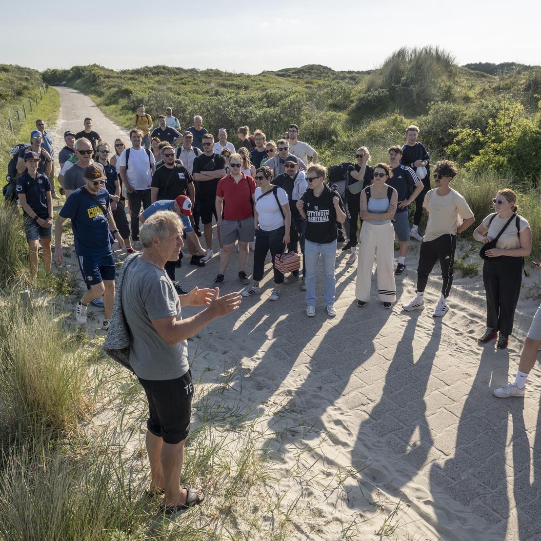 Eine Gruppe von &uuml;ber 30 Personen h&ouml;rt einem Referenten auf einem Sandweg in einer gr&uuml;nen Landschaft zu.