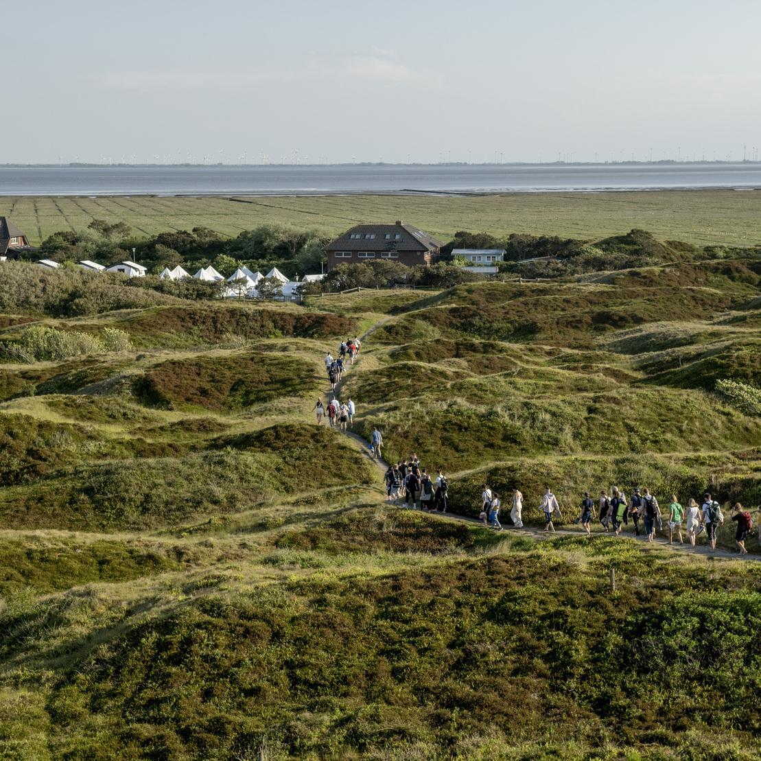 Gruppe von Menschen wandert auf einem Pfad durch h&uuml;gelige Landschaft mit grasbewachsenen D&uuml;nen und H&auml;usern im Hintergrund.
