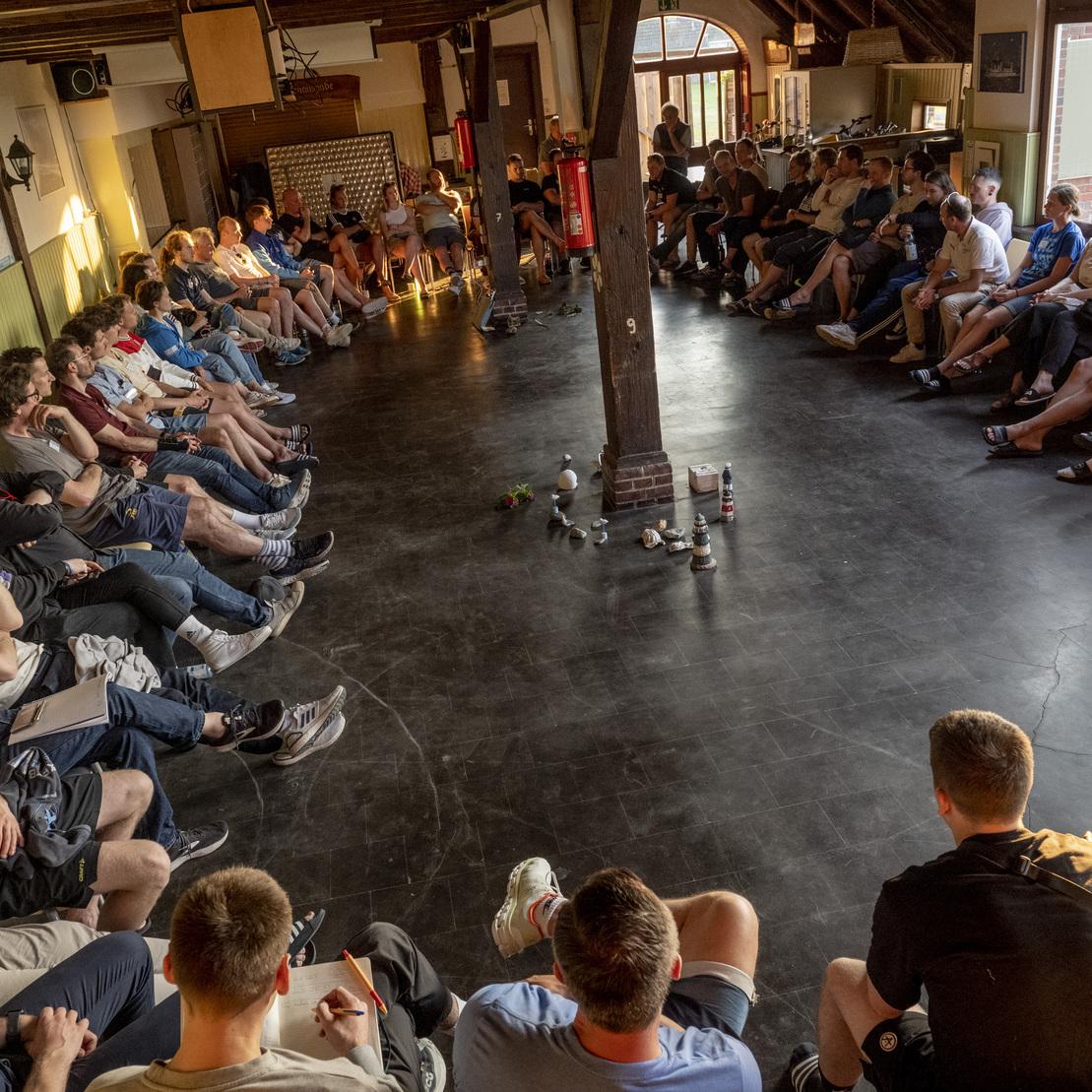 Gruppensitzung mit vielen Menschen, die im Kreis auf St&uuml;hlen sitzen, in einem hellen Raum mit Holzdetails.