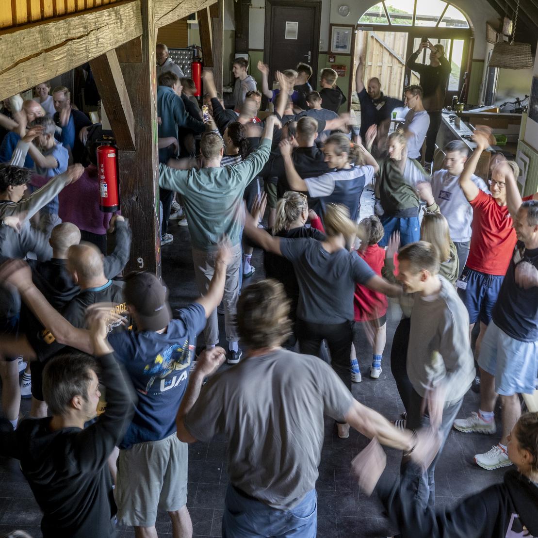 Gruppen von Menschen tanzen und bewegen sich lebhaft in einem Raum mit Holzdecken und gro&szlig;en Fenstern.