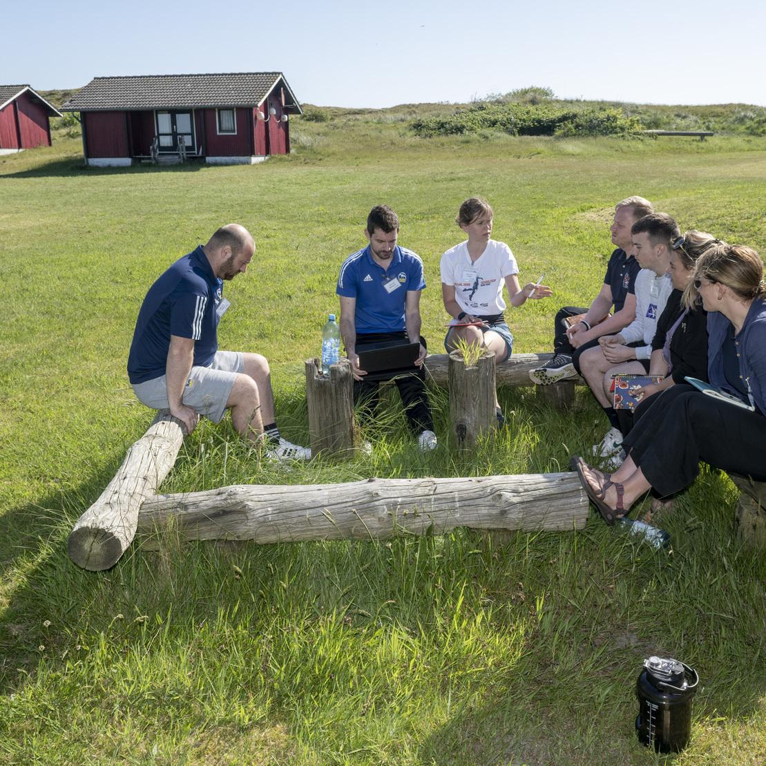 Gruppe von Menschen sitzt im Freien auf Baumst&auml;mmen und bespricht etwas auf einem Laptop. Im Hintergrund rote H&uuml;tten.