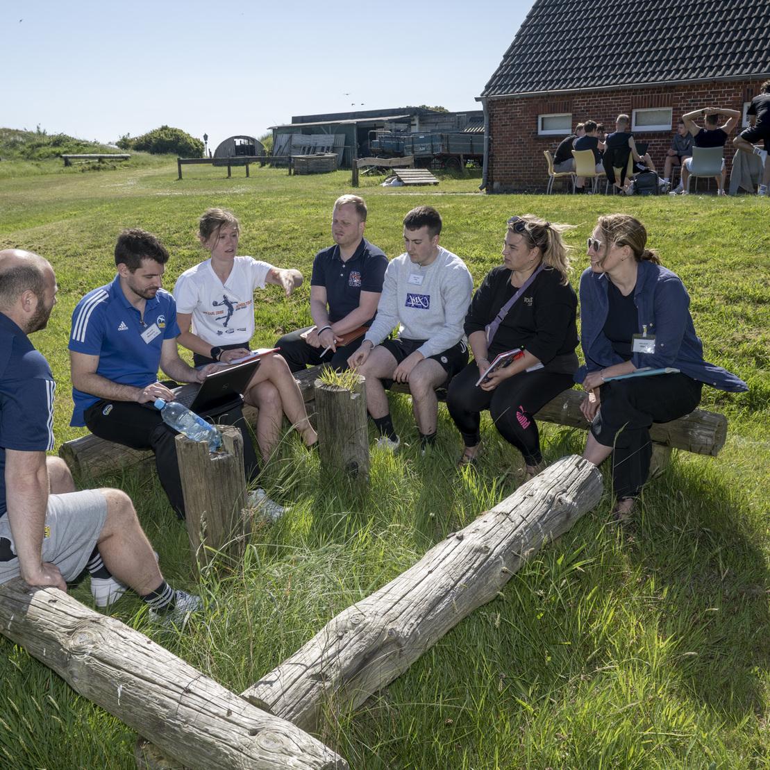 Sechs Personen sitzen im Freien auf Baumst&auml;mmen und diskutieren, w&auml;hrend ein Geb&auml;ude im Hintergrund sichtbar ist.