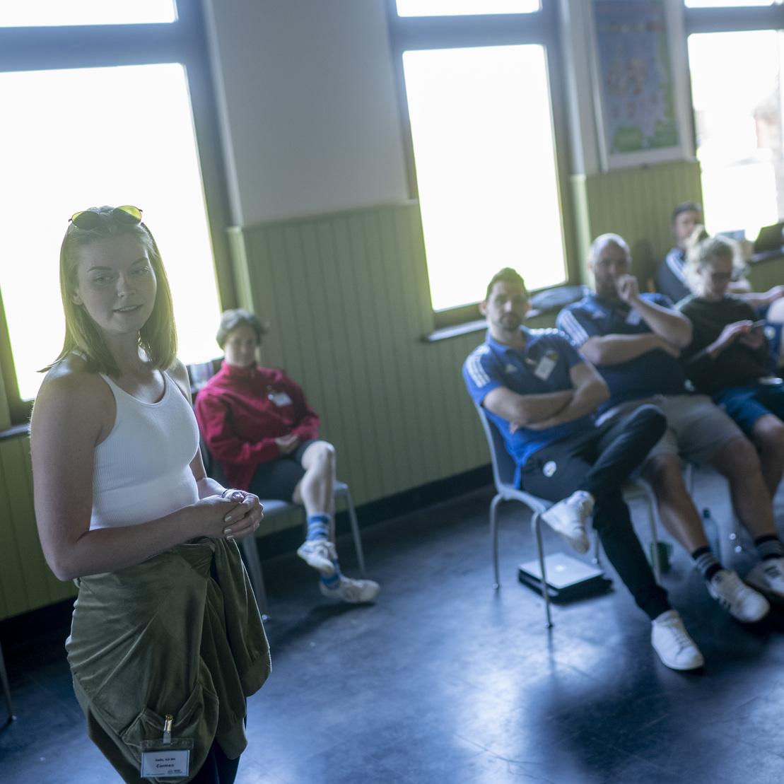 Frau steht an der Wand und spricht zu einer Gruppe von Personen, die in einem Raum auf St&uuml;hlen sitzen.