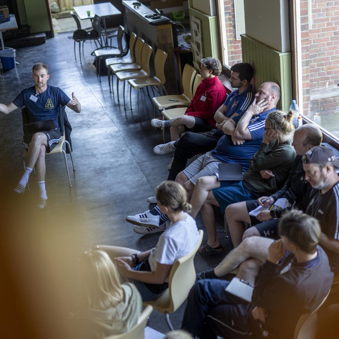 Eine Gruppe von Menschen sitzt auf St&uuml;hlen und h&ouml;rt einem Referenten in einem Raum mit gro&szlig;en Fenstern zu.