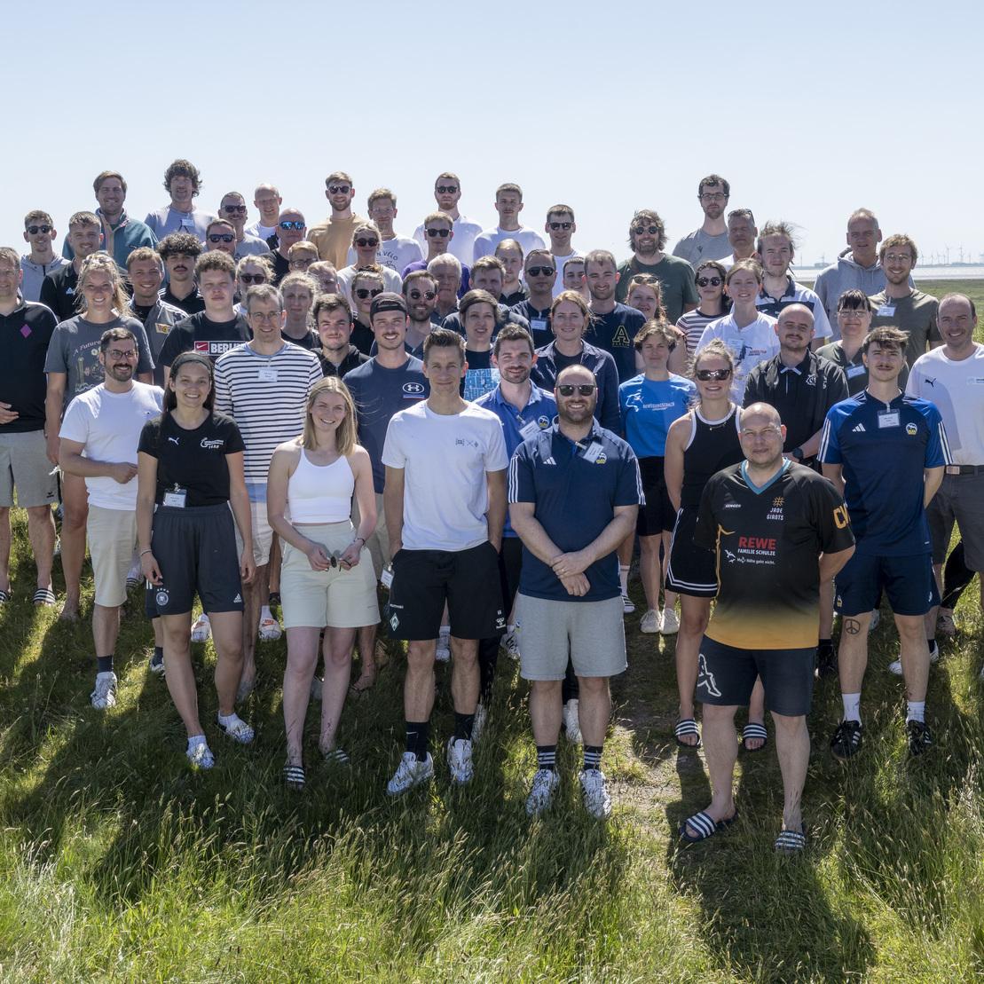 Gruppenfoto von 57 Personen im Freien, vor einer Wiese und Windkraftanlagen im Hintergrund. Alle l&auml;cheln.