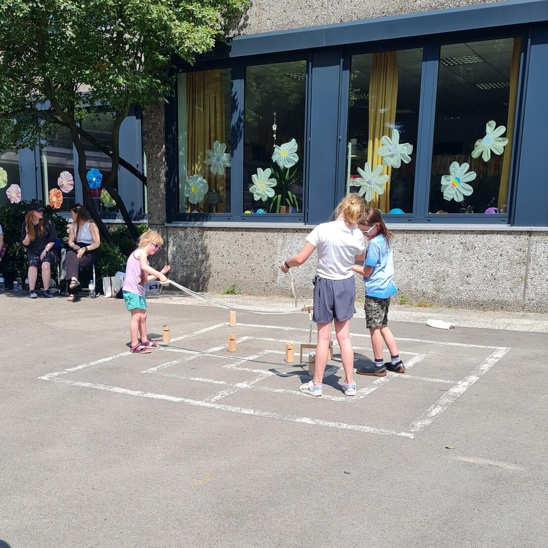 Drei Kinder spielen im Freien mit Holzkl&ouml;tzen und Schn&uuml;ren auf einem gepunkteten Feld vor einem Geb&auml;ude.