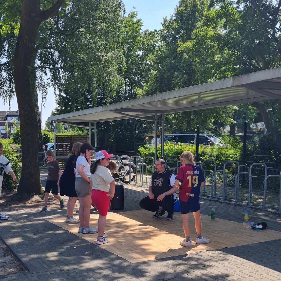Gruppe von Kindern und Jugendlichen auf Holzboden, w&auml;hrend sie einem Trainer im Freien beim Sport zuschauen.