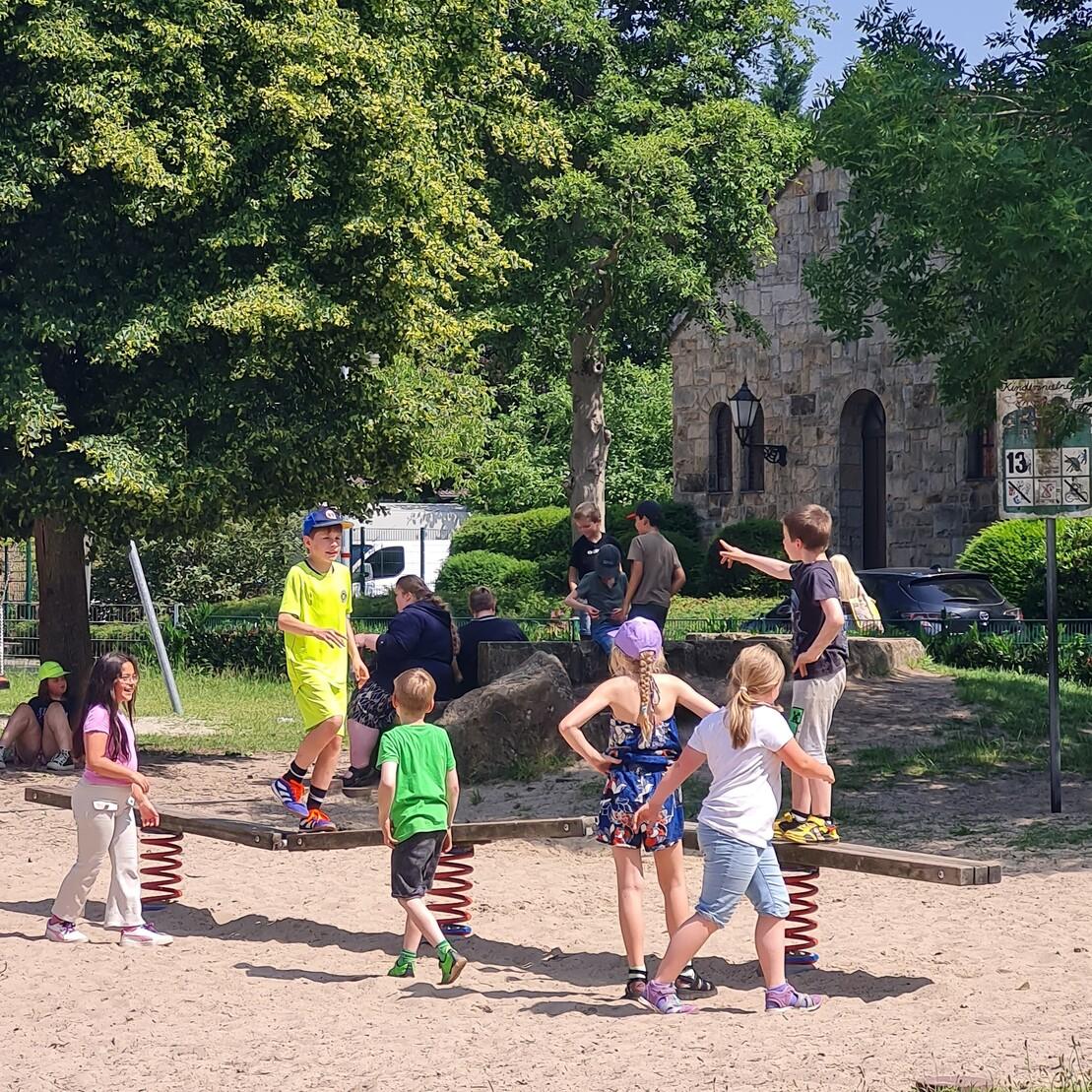 Kinder spielen lebhaft auf einer Wippe im Freien, umgeben von B&auml;umen und einem Steinbau im Hintergrund.