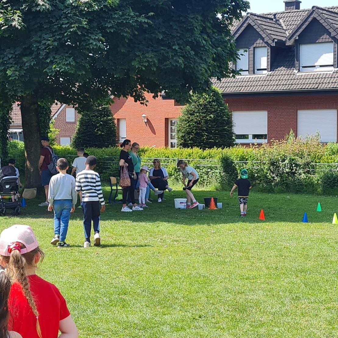 Gruppen von Kindern und Erwachsenen stehen auf einer Wiese, umgeben von bunten H&uuml;tchen und einem Spiel.