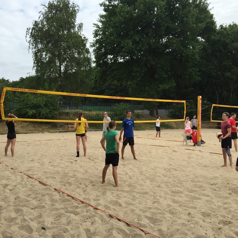 Gruppe von Personen spielt Beachvolleyball auf einem Sandplatz unter einem bew&ouml;lkten Himmel.