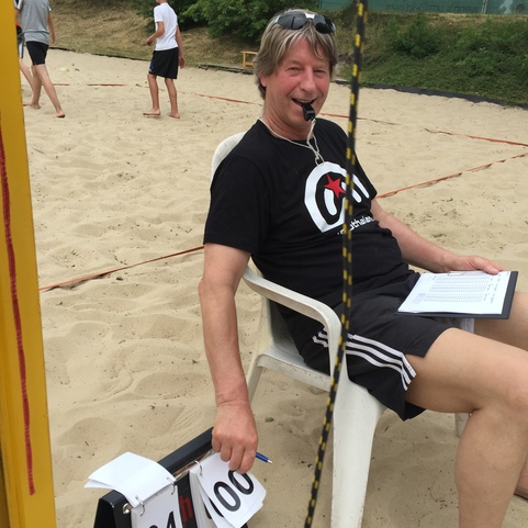 Mann mit Pfeife sitzt auf einem Stuhl im Sand, h&auml;lt ein Punktez&auml;hlger&auml;t und beobachtet ein Beachvolleyballspiel.