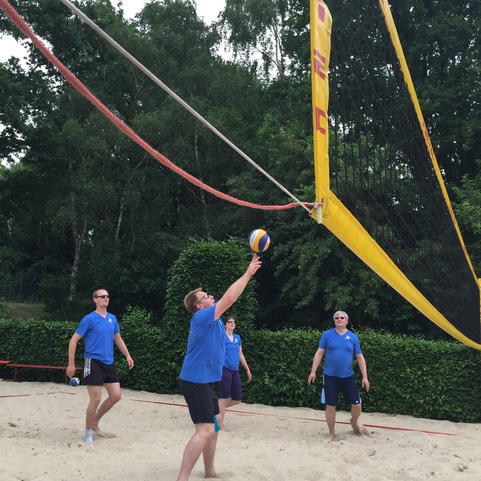 F&uuml;nf Personen in blauen T-Shirts spielen Beachvolleyball am Sandplatz unter B&auml;umen.