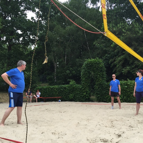 Vier Personen in blauen T-Shirts spielen Beachvolleyball auf einem Sandplatz, umgeben von B&auml;umen und Zuschauern.